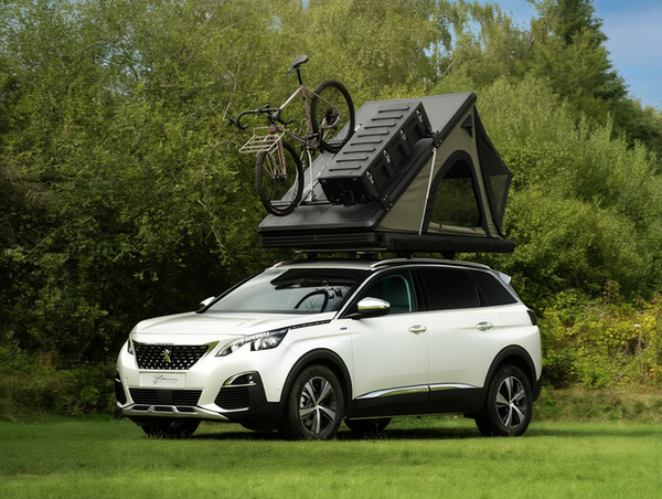 Peugeot 3008 blanc équipé d'une tente de toit rigide noire ouverte et d'un porte-vélo avec un VTT, garé sur une pelouse devant des arbres